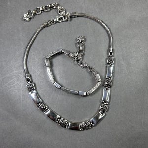 Peut inclure: Un ensemble de bijoux argent&eacute;s comprenant un collier et un bracelet. Le collier pr&eacute;sente une cha&icirc;ne serpent et des maillons rectangulaires d&eacute;coratifs avec des motifs floraux. Le bracelet reprend le design du collier, avec un fermoir assorti. L'ensemble est pr&eacute;sent&eacute; sur une surface grise.
