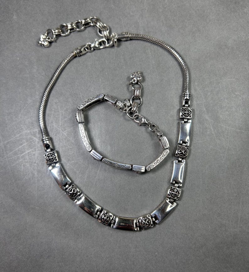 Peut inclure: Un ensemble de bijoux argent&eacute;s comprenant un collier et un bracelet. Le collier pr&eacute;sente une cha&icirc;ne serpent et des maillons rectangulaires d&eacute;coratifs avec des motifs floraux. Le bracelet reprend le design du collier, avec un fermoir assorti. L'ensemble est pr&eacute;sent&eacute; sur une surface grise.