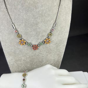 May include: Silver-toned necklace and bracelet set. The necklace features a series of colorful flower-shaped pendants with orange, yellow, green, and red accents. The bracelet, worn on a mannequin hand, mirrors the necklace design.