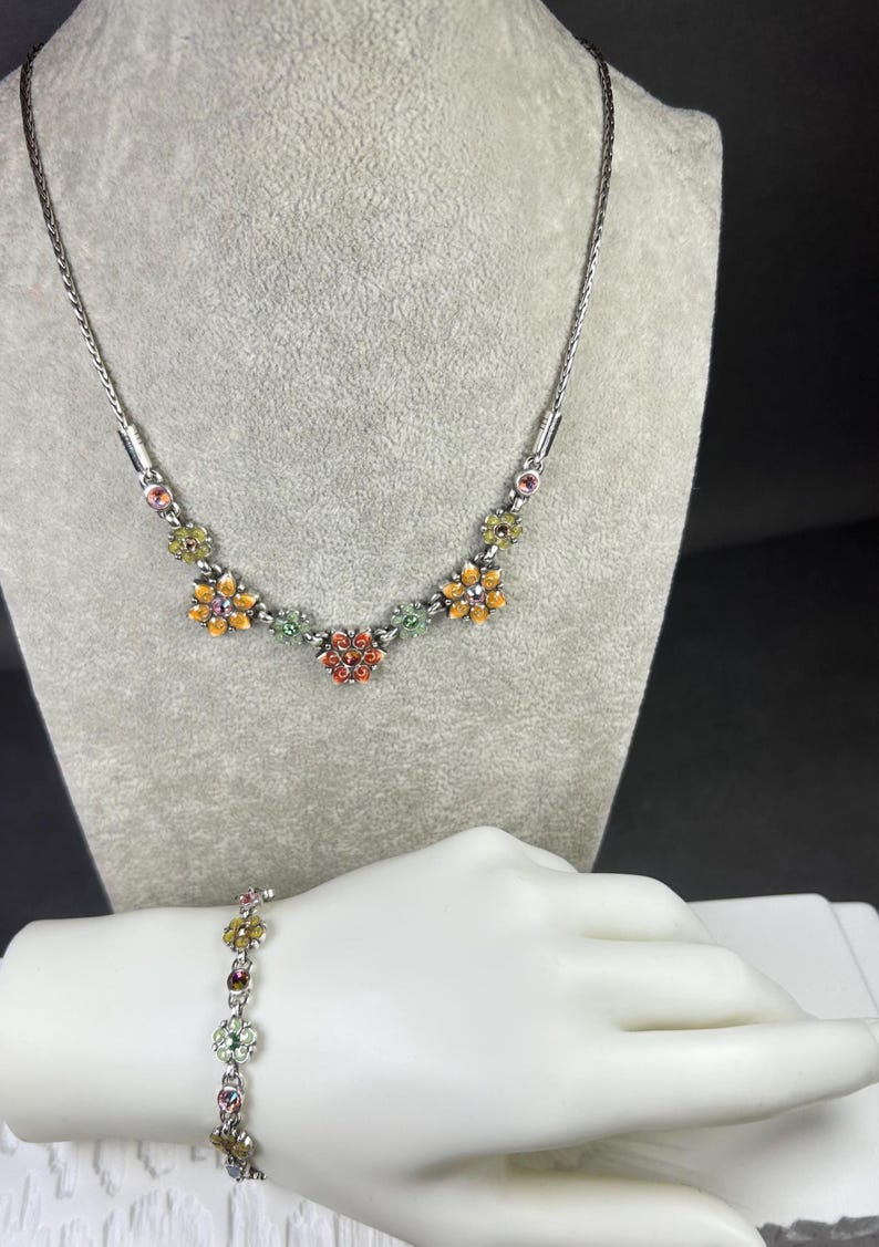 May include: Silver-toned necklace and bracelet set. The necklace features a series of colorful flower-shaped pendants with orange, yellow, green, and red accents. The bracelet, worn on a mannequin hand, mirrors the necklace design.
