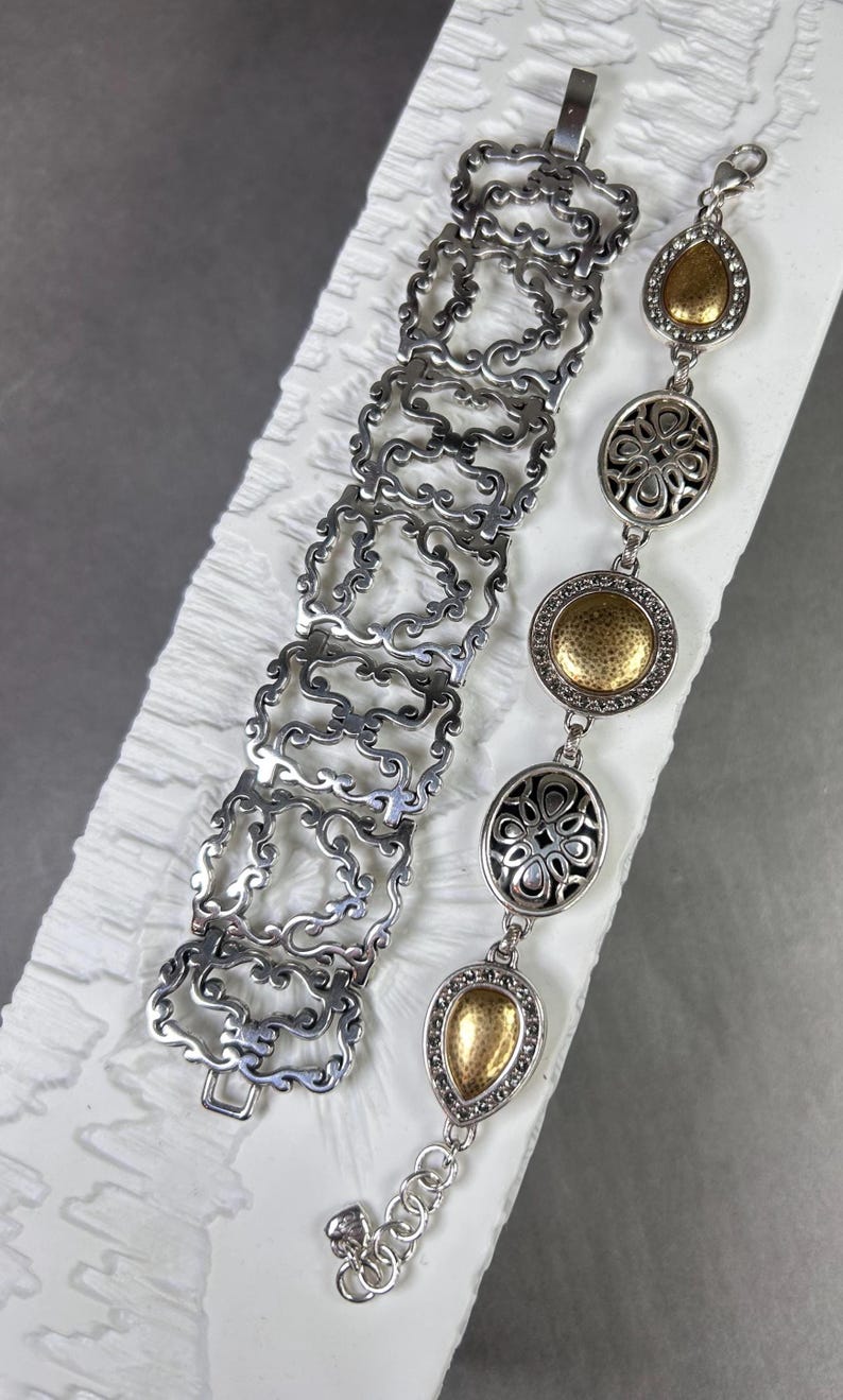 Peut inclure: Deux bracelets argent&eacute;s. L'un pr&eacute;sente un motif ajour&eacute; avec des motifs en volutes. L'autre a des maillons ovales et en forme de goutte d'eau dor&eacute;s et argent&eacute;s. Le bracelet a un fermoir mousqueton.