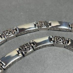 Peut inclure: Gros plan sur deux bracelets en argent. Chaque bracelet est compos&eacute; de maillons rectangulaires polis, reli&eacute;s par de plus petits maillons carr&eacute;s avec des motifs floraux. Les bracelets sont sur une surface grise.