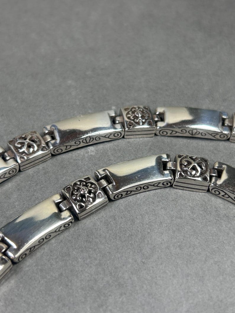 Peut inclure: Gros plan sur deux bracelets en argent. Chaque bracelet est compos&eacute; de maillons rectangulaires polis, reli&eacute;s par de plus petits maillons carr&eacute;s avec des motifs floraux. Les bracelets sont sur une surface grise.