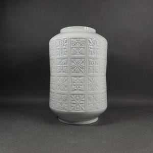 Könnte beinhalten: Eine weiße Keramikvase mit zylindrischem Körper und leicht verjüngtem Hals. Die Außenfläche weist ein sich wiederholendes Gittermuster aus geprägten quadratischen Kacheln auf, die jeweils ein detailliertes Relief aus Blättern und Blüten zeigen.