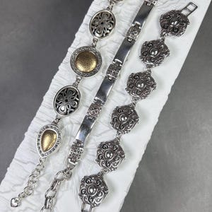 Peut inclure: Trois bracelets argent&eacute;s aux motifs complexes. L'un pr&eacute;sente des formes de goutte et ovales avec des accents dor&eacute;s. Un autre a une s&eacute;rie de maillons rectangulaires connect&eacute;s. Le troisi&egrave;me bracelet a des maillons en forme de fleurs.
