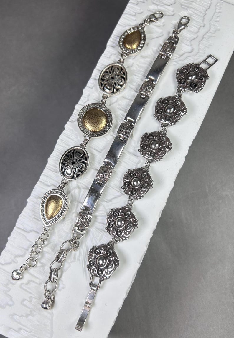 Peut inclure: Trois bracelets argent&eacute;s aux motifs complexes. L'un pr&eacute;sente des formes de goutte et ovales avec des accents dor&eacute;s. Un autre a une s&eacute;rie de maillons rectangulaires connect&eacute;s. Le troisi&egrave;me bracelet a des maillons en forme de fleurs.