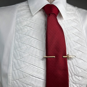 Puede incluir: Un conjunto formal con una camisa blanca con detalles plisados y una corbata rojo oscuro. Una pinza de corbata dorada asegura la corbata, agregando un toque de elegancia. La textura de la camisa y el color de la corbata crean un look clásico.