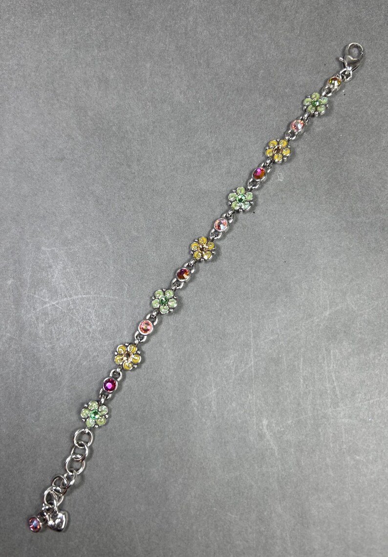 May include: A silver-toned bracelet featuring a series of colorful flower-shaped charms. Each charm is adorned with a cluster of small, sparkling gemstones in shades of pink, yellow, green, and red. The bracelet has a lobster clasp closure.