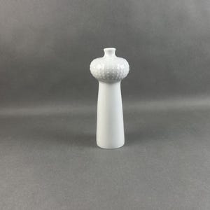Puede incluir: Un salero o pimentero de cerámica blanca con una parte superior texturizada y bulbosa y una base larga y cilíndrica. La parte superior tiene un patrón de pequeños puntos en relieve. El salero está sobre un fondo gris liso.