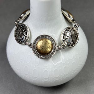 以下が含まれることがあります： 装飾的な円形リンクが付いたシルバーカラーのブレスレット。中央のリンクは、透明な石に囲まれた金色のテクスチャード加工されたセンターを備えています。ブレスレットは、灰色の背景に対して、白いテクスチャード加工された花瓶に表示されています。