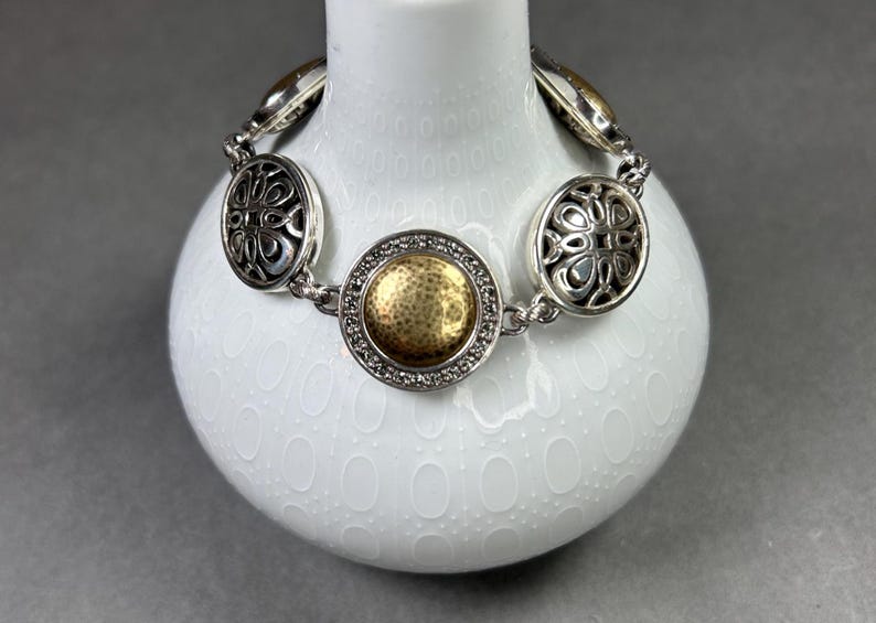 以下が含まれることがあります： 装飾的な円形リンクが付いたシルバーカラーのブレスレット。中央のリンクは、透明な石に囲まれた金色のテクスチャード加工されたセンターを備えています。ブレスレットは、灰色の背景に対して、白いテクスチャード加工された花瓶に表示されています。