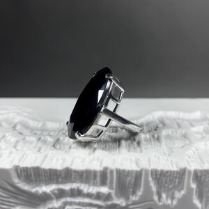 Puede incluir: Un anillo con una gran piedra preciosa oscura de talla marquesa, montada en un engaste de metal plateado. El engaste tiene una estructura abierta tipo jaula con garras visibles que sujetan la piedra facetada, reflejando la luz.