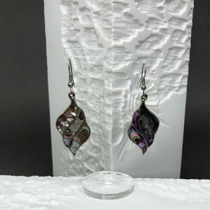 Könnte beinhalten: Ohrringe zum Aufhängen mit einem wirbelnden, muschelartigen Design. Die Ohrringe haben schillernde Abalone-Muscheleinlagen in einem dunklen Metallrahmen. Sie werden auf einem transparenten Acrylständer vor einem strukturierten weißen Hintergrund ausgestellt.