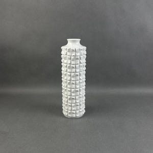 Puede incluir: Un jarrón de cerámica blanco con un patrón geométrico texturizado. El jarrón tiene forma cilíndrica con cuello estrecho y base ancha. La superficie está cubierta por un patrón repetitivo de cuadrados en relieve.
