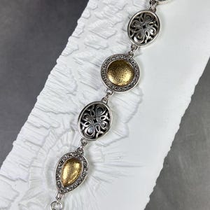 以下が含まれることがあります： ゴールドカラーのアクセントが付いたシルバーカラーのブレスレット。ブレスレットは、複雑なデザインとキラキラしたディテールを備えた、一連の楕円形と涙滴型のリンクが特徴です。留め金はロブスタークロースタイルです。ブレスレットは白い表面に表示されています。