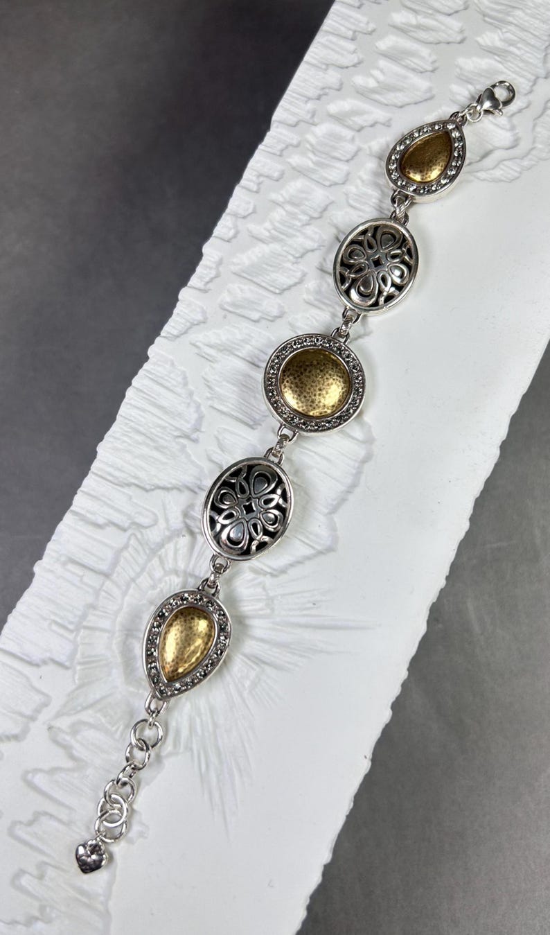 以下が含まれることがあります： ゴールドカラーのアクセントが付いたシルバーカラーのブレスレット。ブレスレットは、複雑なデザインとキラキラしたディテールを備えた、一連の楕円形と涙滴型のリンクが特徴です。留め金はロブスタークロースタイルです。ブレスレットは白い表面に表示されています。