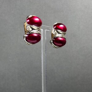 Pode incluir: Um par de brincos vintage de pressão. Cada brinco apresenta duas pedras cabochão ovais, vermelho escuro, emolduradas em metal prateado. Os brincos são exibidos em um suporte de acrílico transparente sobre um fundo cinza.