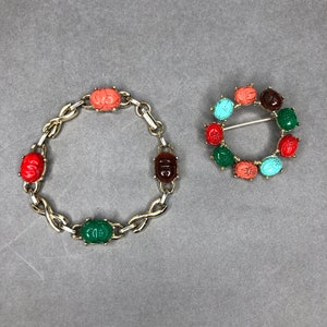 Könnte beinhalten: Ein goldfarbenes Armband- und Broschen-Set. Das Armband verfügt über bunte Skarabäus-Käfer-Anhänger in Rot, Koralle, Grün und Braun. Die Brosche ist ein kreisförmiges Design mit Nadel, ebenfalls mit Skarabäuskäfern in verschiedenen Farben verziert.