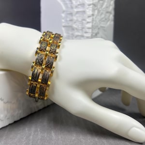 Könnte beinhalten: Ein goldfarbenes Armband mit einem sich wiederholenden rechteckigen Gliederdesign. Jedes Glied weist ein dunkles, geätztes Muster auf. Das Armband wird auf einer weißen Mannequinhand präsentiert, die die komplizierten Details zeigt.