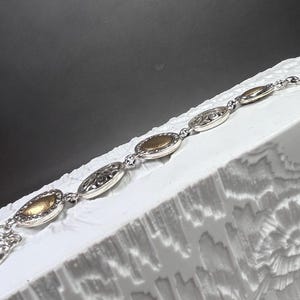 以下が含まれることがあります： 楕円形の金と銀のリンクが付いたシルバーブレスレット。ブレスレットは、彫刻された花の模様が施されたものなど、金と銀の楕円形のリンクが交互に配置されています。ブレスレットは、暗い背景に対して、白くテクスチャ加工された表面に表示されています。
