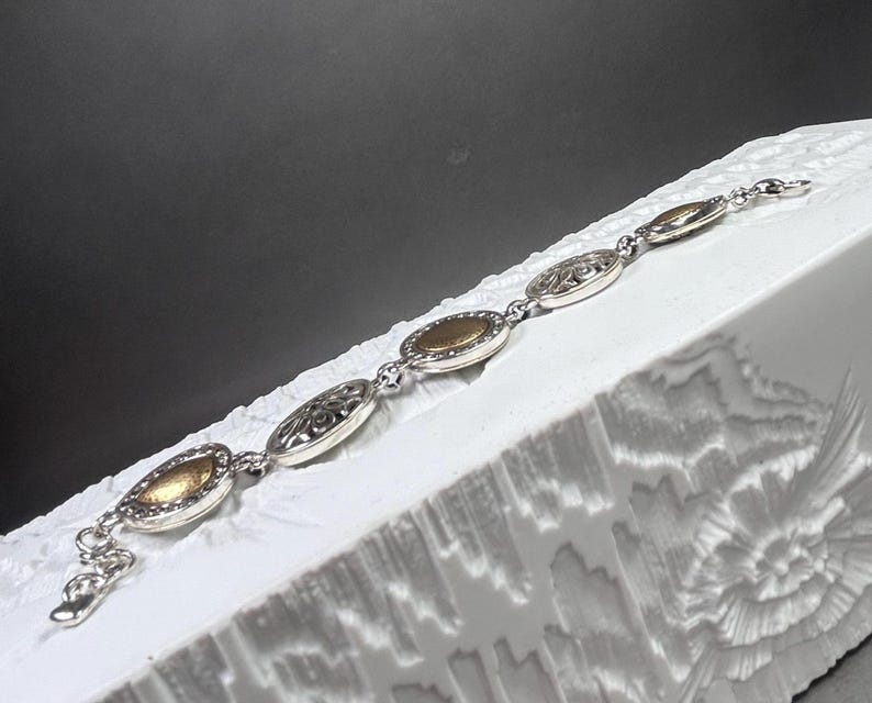 以下が含まれることがあります： 楕円形の金と銀のリンクが付いたシルバーブレスレット。ブレスレットは、彫刻された花の模様が施されたものなど、金と銀の楕円形のリンクが交互に配置されています。ブレスレットは、暗い背景に対して、白くテクスチャ加工された表面に表示されています。