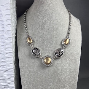 May include: Silver necklace with gold-coloured detailing. The necklace has a chain design with decorative elements, including teardrop and oval shapes. The oval shapes have a black filigree design. The gold elements are surrounded by small, clear stones.