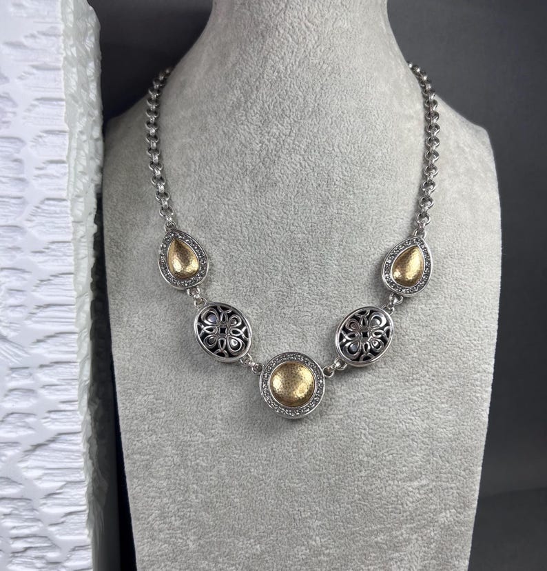 May include: Silver necklace with gold-coloured detailing. The necklace has a chain design with decorative elements, including teardrop and oval shapes. The oval shapes have a black filigree design. The gold elements are surrounded by small, clear stones.