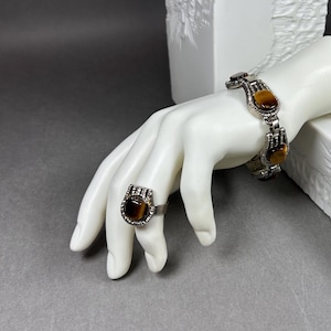 Könnte beinhalten: Ein Ring und ein Gliederarmband aus silberfarbenem Metall, beide mit polierten, rechteckigen Tigerauge-Edelsteinen, die in dekorativen, texturierten Metallrahmen gefasst sind. Die Schmuckstücke werden auf einer weißen Schaufensterpuppenhand präsentiert.