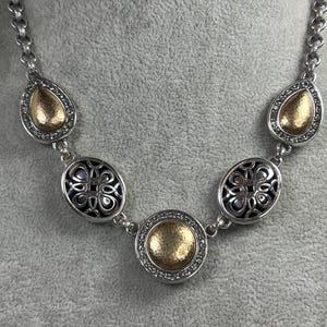 May include: Silver-toned necklace with a chain of oval links. The necklace features decorative elements, including teardrop-shaped and oval pendants with gold-coloured centres and crystal accents. A round pendant is in the centre.