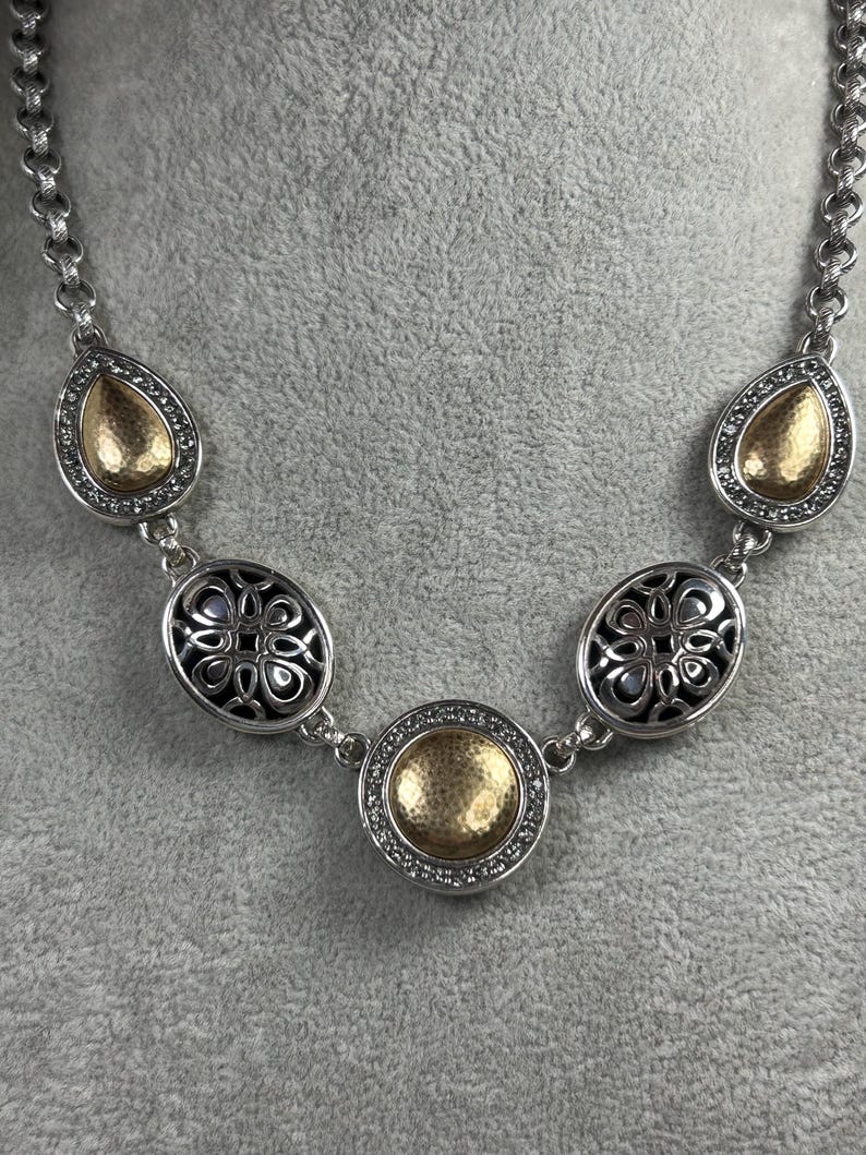 May include: Silver-toned necklace with a chain of oval links. The necklace features decorative elements, including teardrop-shaped and oval pendants with gold-coloured centres and crystal accents. A round pendant is in the centre.