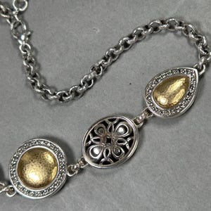 May include: Silver necklace with three decorative pendants. The chain has a lobster clasp. The pendants include a round, an oval, and a teardrop shape, each with a gold-coloured centre and a silver border. The necklace is displayed against a grey background.