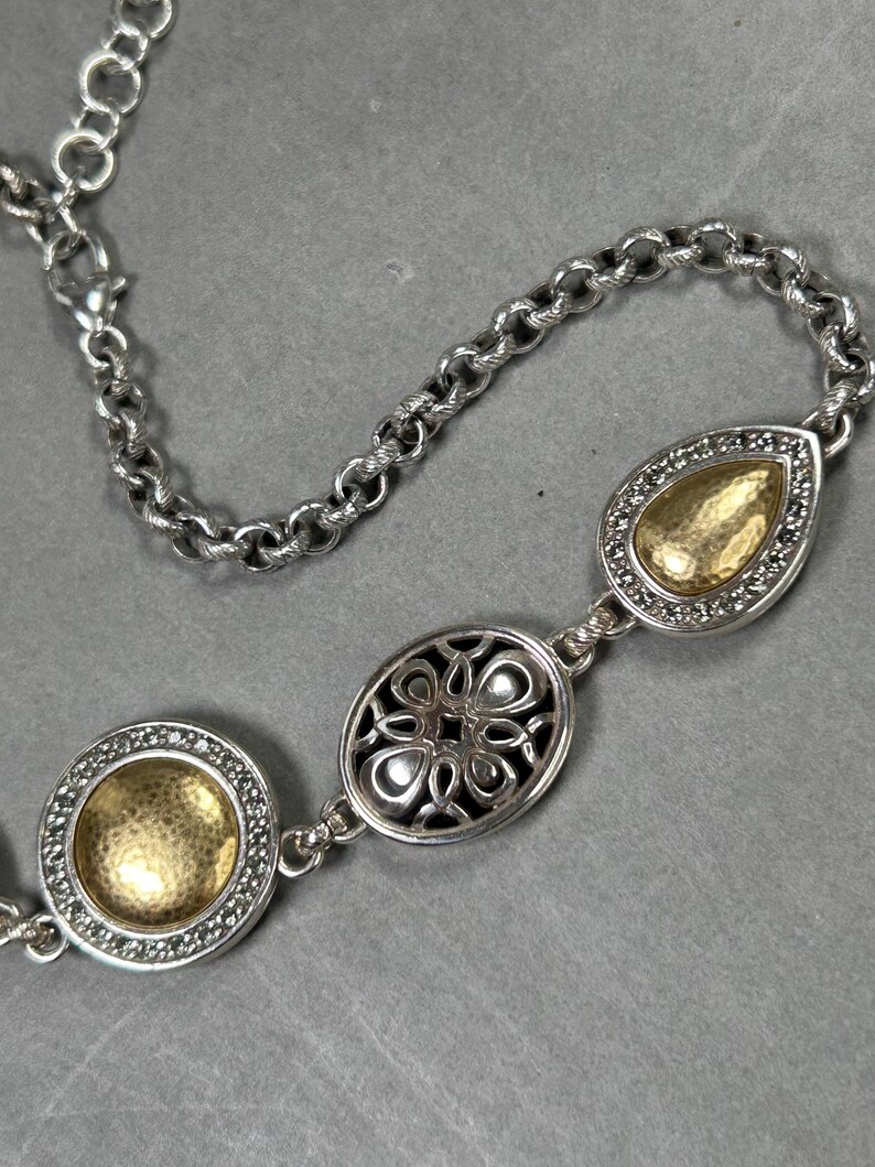 May include: Silver necklace with three decorative pendants. The chain has a lobster clasp. The pendants include a round, an oval, and a teardrop shape, each with a gold-coloured centre and a silver border. The necklace is displayed against a grey background.