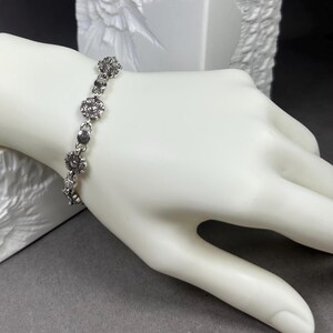 May include: A silver-toned bracelet featuring a floral design. The bracelet is composed of small, round, flower-shaped links connected by smaller, circular links. The bracelet is displayed on a white mannequin hand against a gray background.