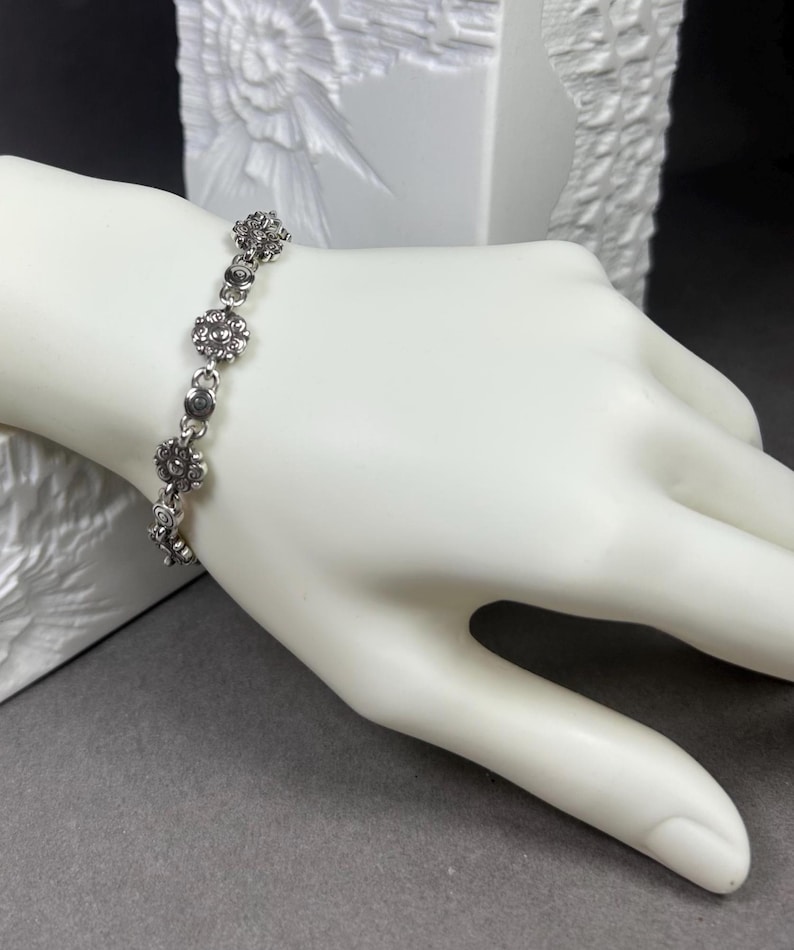 May include: A silver-toned bracelet featuring a floral design. The bracelet is composed of small, round, flower-shaped links connected by smaller, circular links. The bracelet is displayed on a white mannequin hand against a gray background.