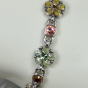 May include: A silver bracelet featuring a floral design. The bracelet has multiple flower charms in yellow, green, and pink, each with a central gemstone. The bracelet is connected by small, round links.