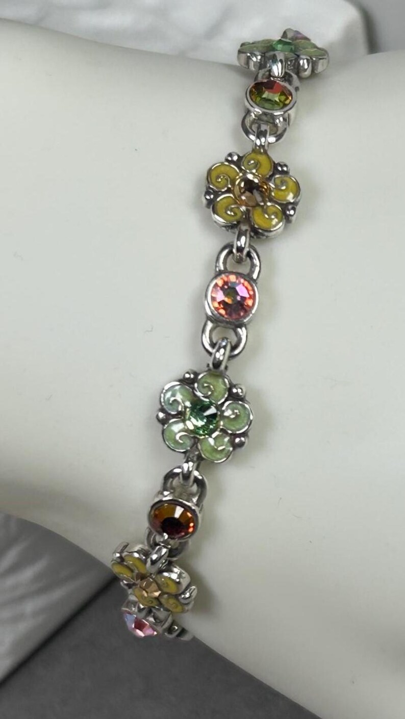 May include: A silver bracelet featuring a floral design. The bracelet has multiple flower charms in yellow, green, and pink, each with a central gemstone. The bracelet is connected by small, round links.