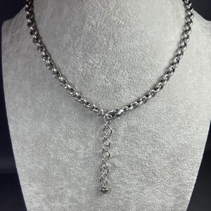 May include: A silver necklace with a chain-link design. The necklace is displayed on a grey mannequin. The chain is made up of circular links, with an adjustable extension and a small charm at the end.