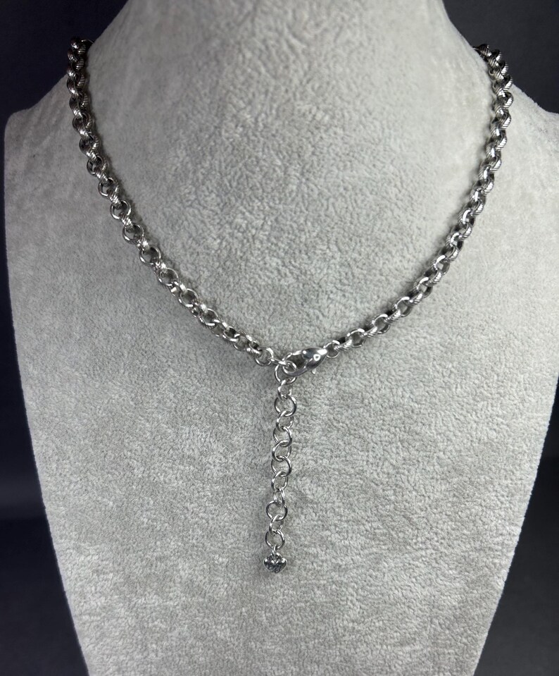 May include: A silver necklace with a chain-link design. The necklace is displayed on a grey mannequin. The chain is made up of circular links, with an adjustable extension and a small charm at the end.