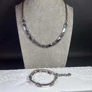 Peut inclure: Un ensemble collier et bracelet argent&eacute;. Le collier est pr&eacute;sent&eacute; sur un mannequin gris, et le bracelet repose sur une surface blanche. Les deux pi&egrave;ces pr&eacute;sentent des maillons rectangulaires avec des accents floraux d&eacute;coratifs.