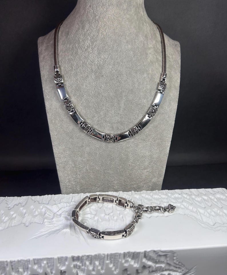 Peut inclure: Un ensemble collier et bracelet argent&eacute;. Le collier est pr&eacute;sent&eacute; sur un mannequin gris, et le bracelet repose sur une surface blanche. Les deux pi&egrave;ces pr&eacute;sentent des maillons rectangulaires avec des accents floraux d&eacute;coratifs.