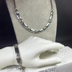Peut inclure: Un ensemble collier et bracelet argent&eacute;. Le collier pr&eacute;sente des maillons rectangulaires avec des d&eacute;tails d&eacute;coratifs, et le bracelet reprend ce design. Les bijoux sont pr&eacute;sent&eacute;s sur un mannequin gris et une main blanche.