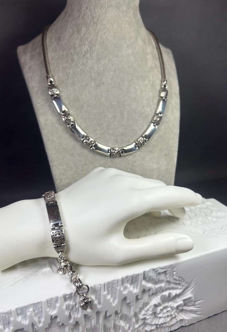 Peut inclure: Un ensemble collier et bracelet argent&eacute;. Le collier pr&eacute;sente des maillons rectangulaires avec des d&eacute;tails d&eacute;coratifs, et le bracelet reprend ce design. Les bijoux sont pr&eacute;sent&eacute;s sur un mannequin gris et une main blanche.