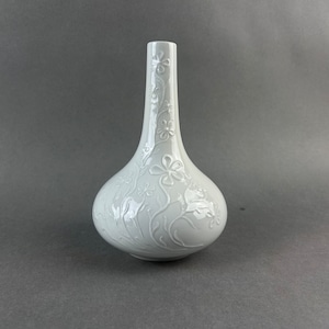 Puede incluir: Un jarrón de cerámica blanca con base bulbosa y cuello esbelto. El jarrón está adornado con un relieve floral de flores y vides. El jarrón mide aproximadamente 25 cm de altura.