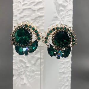 Könnte beinhalten: Ein Paar Vintage-Ohrringe mit smaragdgrünen Strasssteinen. Jeder Ohrring hat einen großen, facettierten grünen Stein, der von kleineren grünen Steinen und einem goldfarbenen Metallrahmen umgeben ist. Die Ohrringe haben ein florales Design.