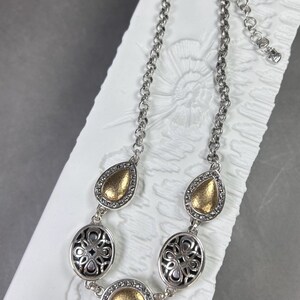 May include: Silver necklace with gold-coloured details. The necklace has a chain with teardrop and oval-shaped pendants. The teardrop pendants have a gold-coloured centre, while the oval pendants have a black filigree design.