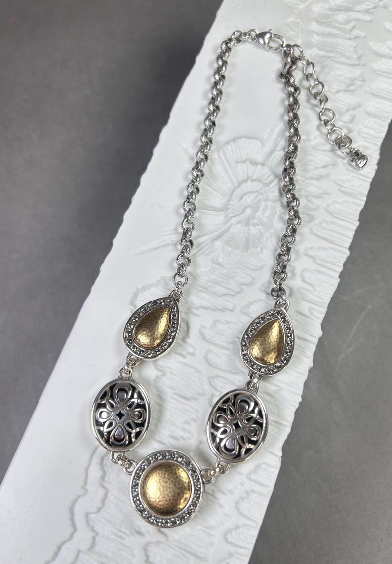 May include: Silver necklace with gold-coloured details. The necklace has a chain with teardrop and oval-shaped pendants. The teardrop pendants have a gold-coloured centre, while the oval pendants have a black filigree design.