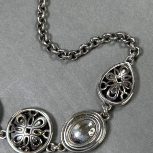May include: Silver necklace featuring a chain and three decorative pendants. The pendants include a circular design with a floral pattern, an oval-shaped stone, and a teardrop-shaped pendant with a floral design. The necklace is displayed on a grey surface.