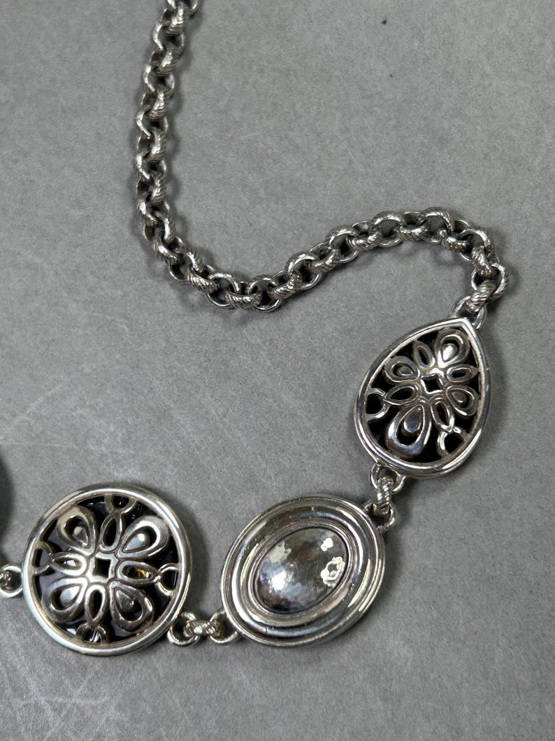 May include: Silver necklace featuring a chain and three decorative pendants. The pendants include a circular design with a floral pattern, an oval-shaped stone, and a teardrop-shaped pendant with a floral design. The necklace is displayed on a grey surface.