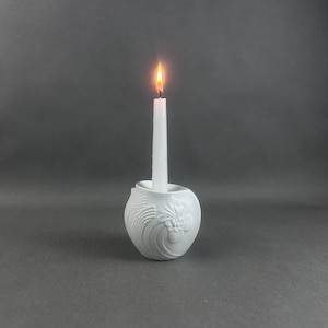 May include: A white ceramic candle holder with a floral and swirl design, holding a lit white candle. The candle flame is bright orange and yellow. The candle holder is round and sits on a gray surface.