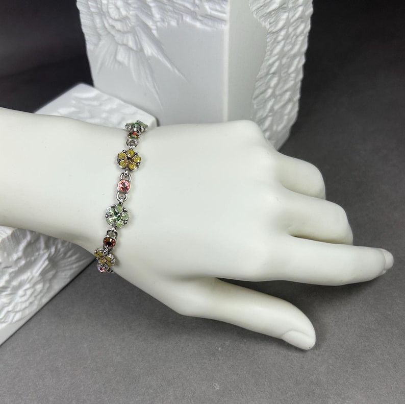 May include: A silver bracelet with flower-shaped charms. Each charm features a cluster of colorful gemstones in shades of yellow, pink, green, and brown. The bracelet is displayed on a white mannequin hand.